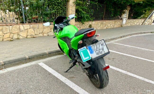 Detalle de la parte trasera de una motocicleta con la placa 'L' de conductor novel correctamente instalada mediante un soporte junto a la matrícula.